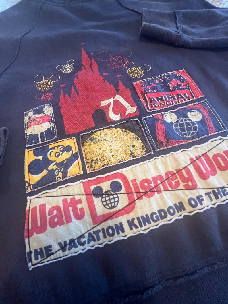 May include: A dark navy sweatshirt featuring a collage of patches with Disney themes. The design includes a red castle, Mickey Mouse, the text Walt Disney World, The Vacation Kingdom of the World, and Animal Kingdom, all stitched onto the fabric.