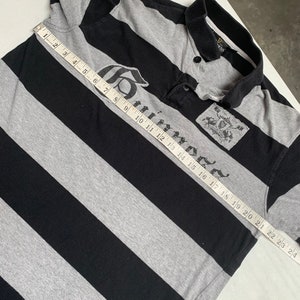 May include: A black and gray striped polo shirt with a large "Guinness" logo printed on the front. The shirt has a button-up collar and short sleeves. The shirt is measured with a tape measure, showing a length of 24 inches.