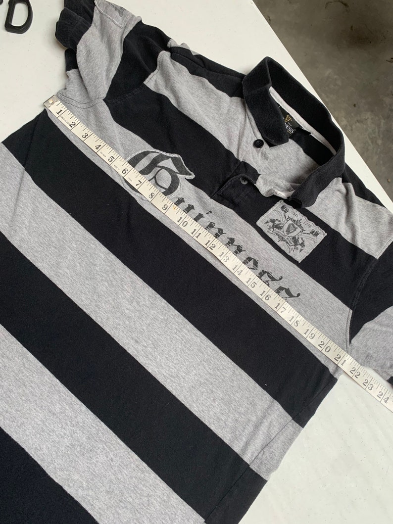 May include: A black and gray striped polo shirt with a large "Guinness" logo printed on the front. The shirt has a button-up collar and short sleeves. The shirt is measured with a tape measure, showing a length of 24 inches.