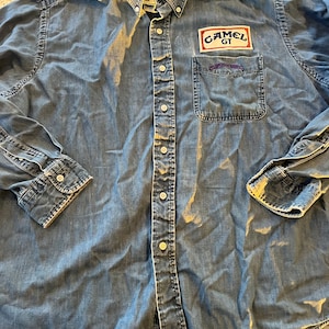 Peut inclure: Une chemise en jean bleu à manches longues avec un bouton sur la poitrine gauche qui dit "Camel GT".