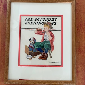 Puede incluir: Un bordado de punto de cruz enmarcado de una ilustración de Norman Rockwell de The Saturday Evening Post, fechado el 10 de marzo de 1923. La ilustración representa a un niño pequeño sentado en un taburete, sosteniendo un vaso de leche y mirando a un perro sentado en una caja de madera.