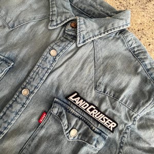 Könnte beinhalten: Hellblaues Jeanshemd mit Knopfleiste, zwei Brusttaschen und Kragen. Mit einem "LAND CRUISER" Aufnäher und einem roten "Levi's" Etikett. Das Hemd hat silberne Knöpfe und einen klassischen Western-Stil.