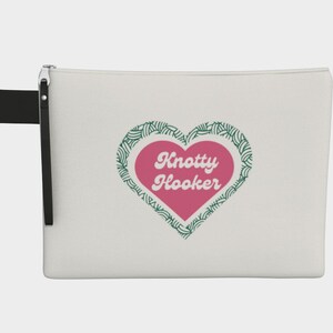 May include: A white zippered pouch with a black wrist strap. The pouch features a pink heart with the words "Knotty Hooker" in white, surrounded by a green leaf design. The pouch is designed for storing craft supplies.