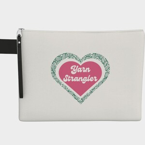 May include: A cream-colored zippered pouch with a black wrist strap. The pouch features a pink heart with the words "Yarn Strangler" in white, surrounded by a green leaf design. The pouch is designed for yarn enthusiasts.