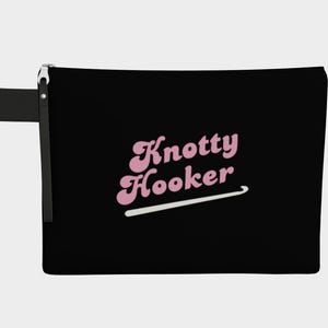 May include: Black zippered pouch with the text "Knotty Hooker" in pink script and a white crochet hook graphic. The pouch has a black wrist strap attached to the zipper.