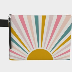 May include: A rectangular zippered pouch with a colorful sunburst design. The design features a yellow sun at the bottom with radiating stripes in pink, teal, and gold against a white background. A black wrist strap is attached.