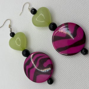 May include: A pair of dangle earrings with black and green beads. The earrings feature a pink and black zebra print circle and a light green heart.