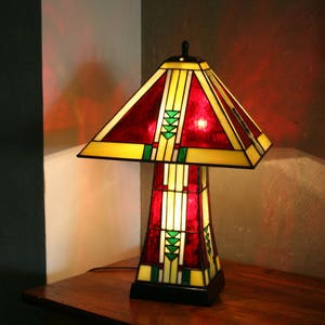 May include: A stained glass table lamp with a red, yellow, and green geometric design. The lamp is turned on and casting a warm glow.