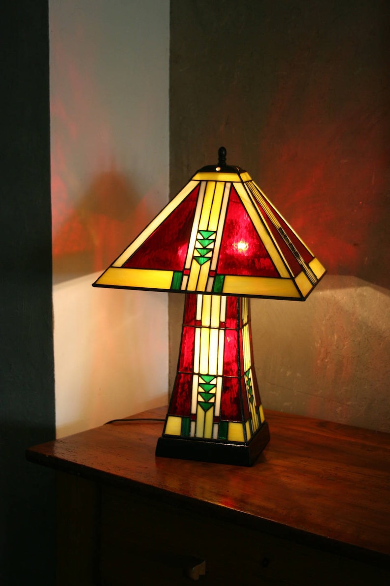 May include: A stained glass table lamp with a red, yellow, and green geometric design. The lamp is turned on and casting a warm glow.