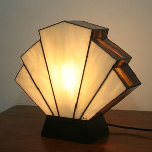 May include: A white and brown stained glass table lamp with a fan-shaped shade. The lamp is switched on and the light is shining through the stained glass.