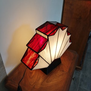 Op de afbeelding: Een tafellamp van gekleurd glas met een geometrisch ontwerp in rood en wit. De lamp is aan en verlicht de kamer.