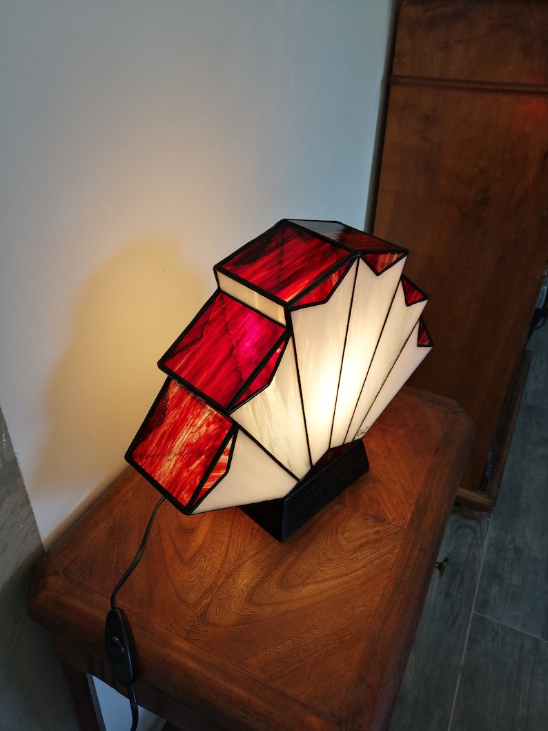 Op de afbeelding: Een tafellamp van gekleurd glas met een geometrisch ontwerp in rood en wit. De lamp is aan en verlicht de kamer.