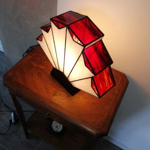 Op de afbeelding: Een rood-witte Tiffany-lamp in de vorm van een waaier. De lamp staat op een houten tafel.