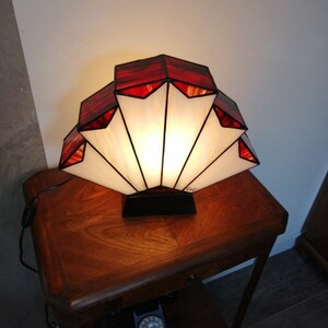 Op de afbeelding: Een lamp van gekleurd glas met een waaiervormig ontwerp. De lamp heeft rode en witte glaspanelen en wordt verlicht met een warm wit licht. De lamp staat op een houten tafel.