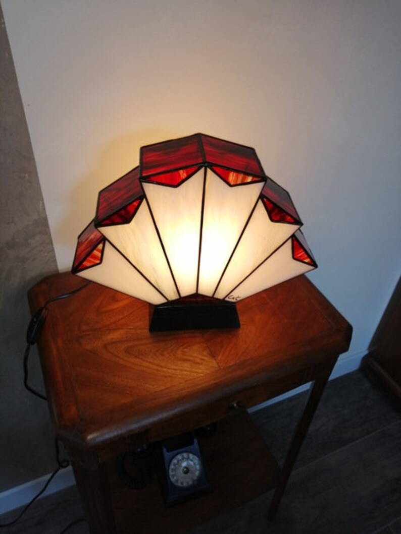 Op de afbeelding: Een lamp van gekleurd glas met een waaiervormig ontwerp. De lamp heeft rode en witte glaspanelen en wordt verlicht met een warm wit licht. De lamp staat op een houten tafel.