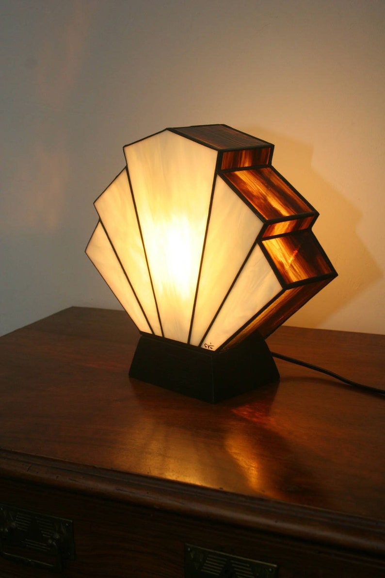 May include: A stained glass lamp with a fan-shaped shade in white and brown. The lamp is on a wooden table with drawers.