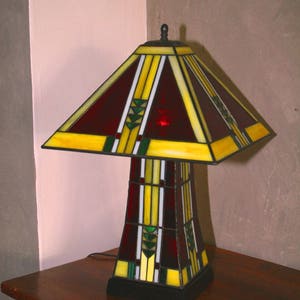May include: A stained glass table lamp with a red, yellow, and green geometric design. The lamp has a square base and a tall, rectangular shade.