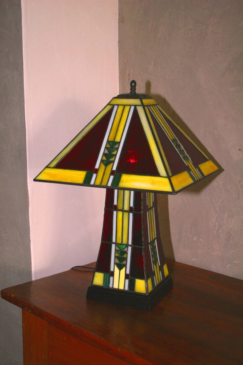 May include: A stained glass table lamp with a red, yellow, and green geometric design. The lamp has a square base and a tall, rectangular shade.