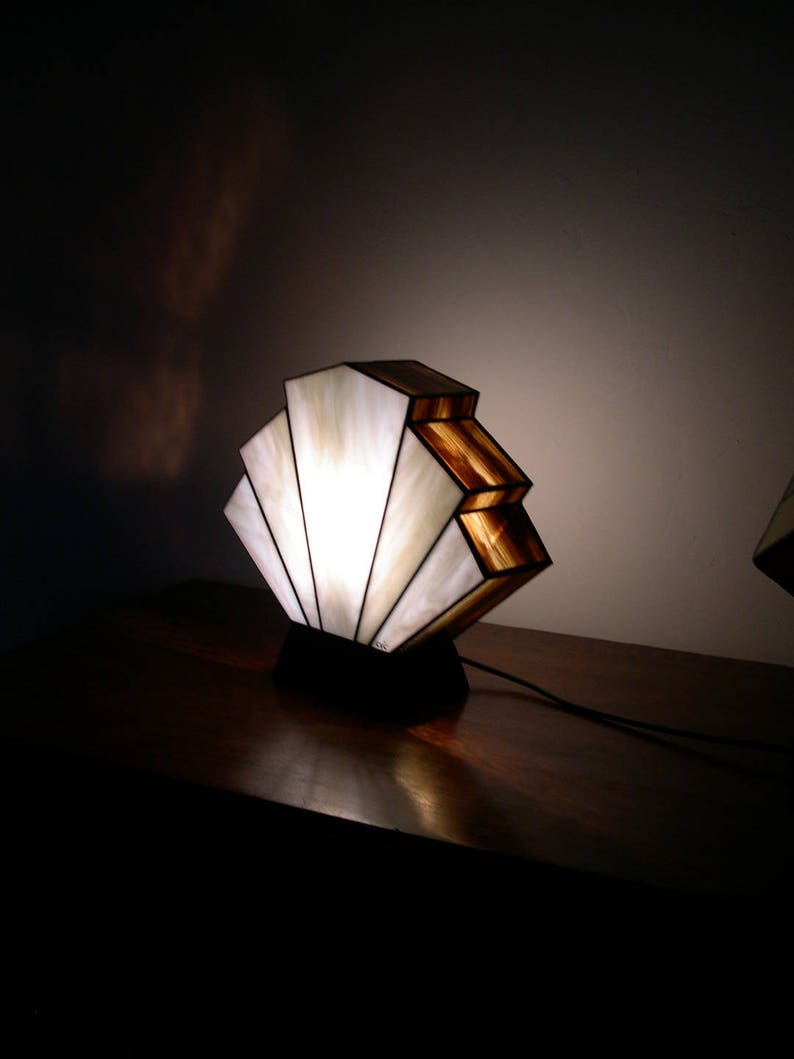 May include: A white and brown stained glass lamp with a fan-shaped design. The lamp is on a dark wooden surface and is lit.