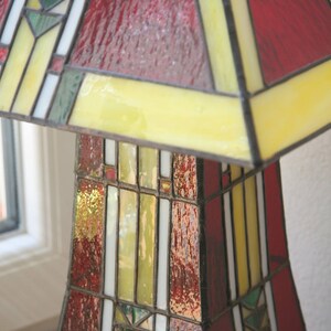May include: A stained glass lamp with a geometric design in shades of red, yellow, and green. The lamp has a square base and a triangular shade.