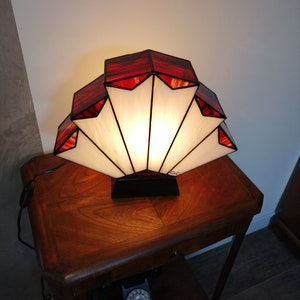 Op de afbeelding: Een rood-witte Tiffany-lamp met een waaiervormig ontwerp. De lamp staat op een houten tafel met een vintage draaischijftelefoon.