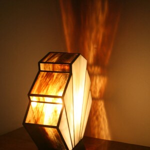 May include: A stained glass table lamp with a geometric design. The lamp has a brown and white colour scheme and is lit with a warm white light bulb. The lamp is sitting on a wooden table.