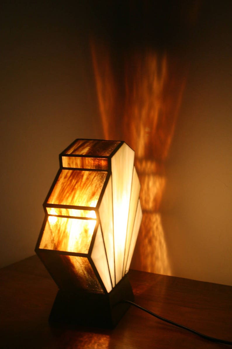 May include: A stained glass table lamp with a geometric design. The lamp has a brown and white colour scheme and is lit with a warm white light bulb. The lamp is sitting on a wooden table.