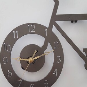 May include: A gray metal wall clock shaped like a bicycle with gold-colored hands. The clock face has black numbers 1 through 12.