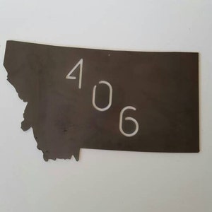 May include: A metal cutout of the state of Montana with the number 406 cut out in white. The cutout is a dark brown color.