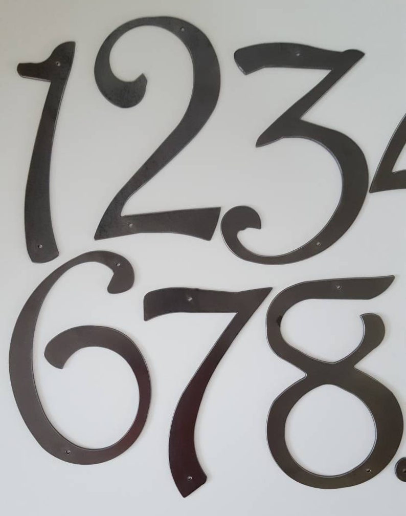 May include: A set of black metal house numbers, 1, 2, 3, 6, 7, and 8, with a decorative, cursive font. The numbers are individually cut and have a slightly distressed finish.