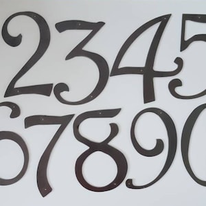 May include: Set of ten black metal house numbers with a decorative, cursive font. The numbers are 1, 2, 3, 4, 5, 6, 7, 8, 9, and 0.