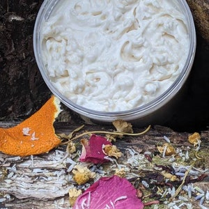 May include: A clear plastic jar filled with white, whipped cream-like product. The jar sits on a wooden surface, surrounded by dried flowers, a pink petal, and a piece of orange peel. The product appears to be a skincare item.