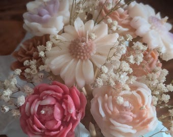 Op de afbeelding: Een boeket van bloemvormige kaarsen in verschillende kleuren, waaronder roze, perzik en wit. De kaarsen zijn gerangschikt met delicate witte vulbloemen, waardoor een bloemstuk ontstaat. De kaarsen worden gepresenteerd op lichtblauw papier.