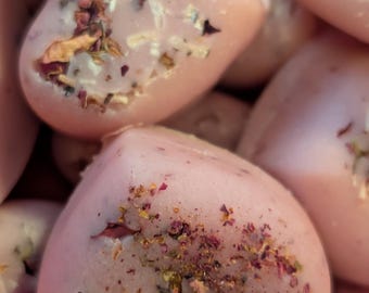 Op de afbeelding: Close-up van verschillende hartvormige, lichtroze wax melts. Elke melt is versierd met gedroogde bloemblaadjes en kruiden. De melts hebben een gladde, glanzende uitstraling en de achtergrond is wazig.