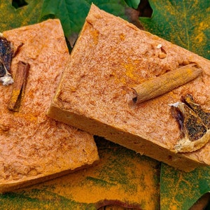 Op de afbeelding: Twee vierkante, lichtbruine bakproducten, mogelijk zeep of een dessert, worden gepresenteerd op een groen blad. Elk is gegarneerd met een gedroogde sinaasappelschijf en een kaneelstokje. Geel poeder is rond de bakproducten gestrooid.