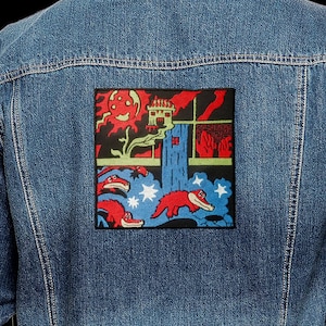 May include: A blue denim jacket with a colourful embroidered patch on the back. The patch features a red and yellow sun, a blue building, a red and yellow waterfall, and red and yellow alligators swimming in a blue river.