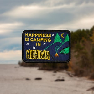 May include: A blue embroidered patch with yellow text that reads "Happiness is camping in Michigan". The patch features a green tent, a campfire, and a crescent moon.