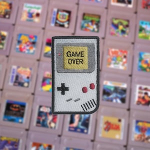 May include: An embroidered patch of a handheld video game console in grey and white, with the words "GAME OVER" displayed on the screen.