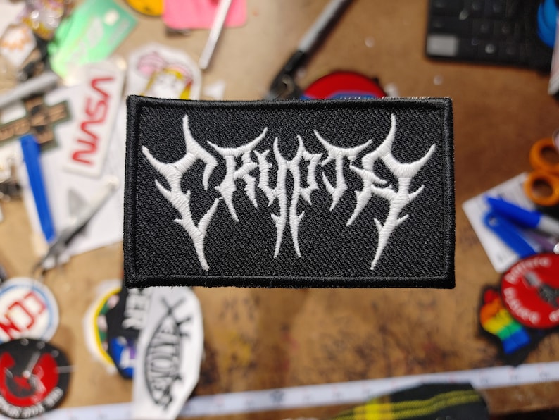 以下が含まれることがあります： 白い文字「CRYPTIC」がスタイリッシュなフォントで刺繍された黒いパッチ