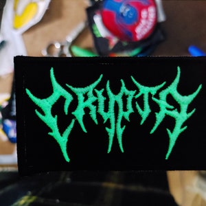 以下が含まれることがあります： 黒い背景にスタイリッシュなフォントで「CRYPTIC」という言葉が書かれた緑色の刺繍パッチ