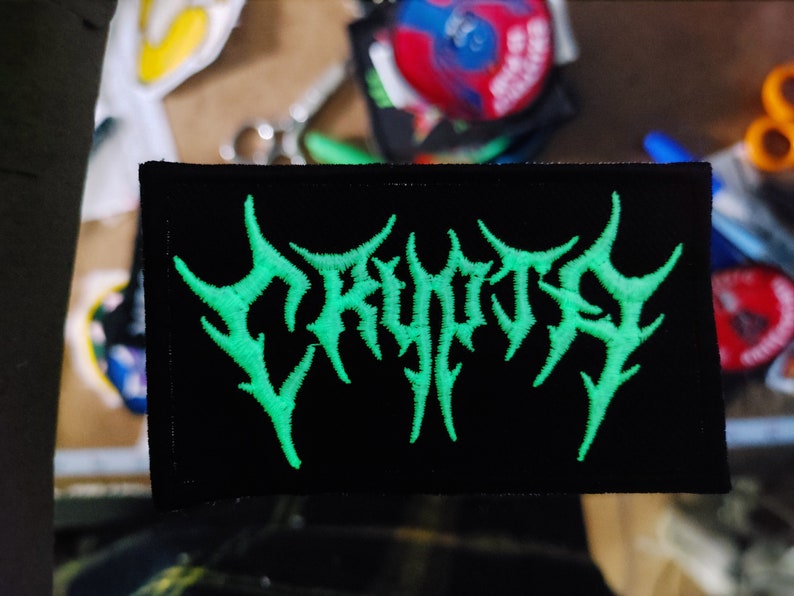 以下が含まれることがあります： 黒い背景にスタイリッシュなフォントで「CRYPTIC」という言葉が書かれた緑色の刺繍パッチ
