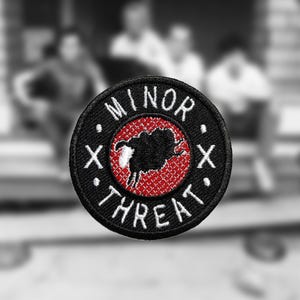 Op de afbeelding: Zwart-witte geborduurde patch met een rode achtergrond. De patch heeft een zwart schaap met een wit gezicht en de tekst "MINOR THREAT" in wit.