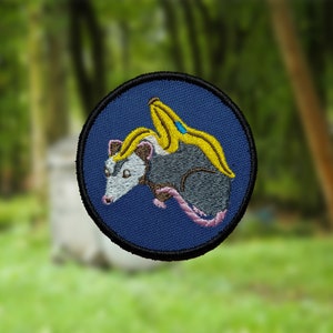 May include: Embroidered patch featuring a gray opossum with a yellow banana on its back, against a dark blue background.