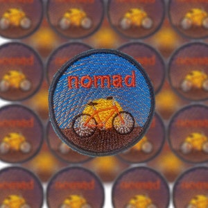 Peut inclure: Un patch brodé bleu et marron avec le mot "Nomad" en lettres rouges et une silhouette de vélo avec un cadre jaune.