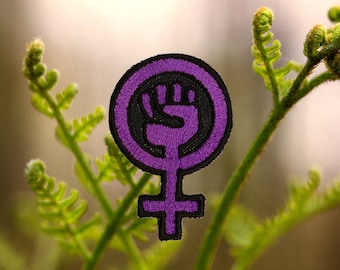 May include: Purple feminist symbol with a raised fist in the center. The symbol is a circle with a cross underneath. The fist is black and the circle and cross are purple.