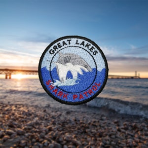 以下が含まれることがあります： 「Great Lakes Shark Patrol」という文字と白いサメのイラストが刺繍された青と白のワッペンです。