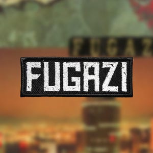 Puede incluir: Parche bordado en blanco y negro con la palabra "FUGAZI" en letras blancas.