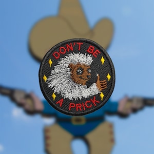 Peut inclure: Un écusson brodé noir avec un porc-épic blanc donnant un pouce en l'air. Le texte "DON'T BE A PRICK" est en rouge.