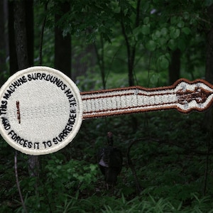 Peut inclure: Écusson brodé en forme de banjo. La tête circulaire du banjo porte l'inscription en noir : « THIS MACHINE SURROUNDS HATE AND FORCES IT TO SURRENDER ». Le manche du banjo est marron et blanc.