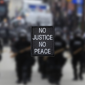 May include: A black patch with white text that reads "NO JUSTICE NO PEACE". The patch is in focus, while the background is blurred and shows a group of people in black uniforms.